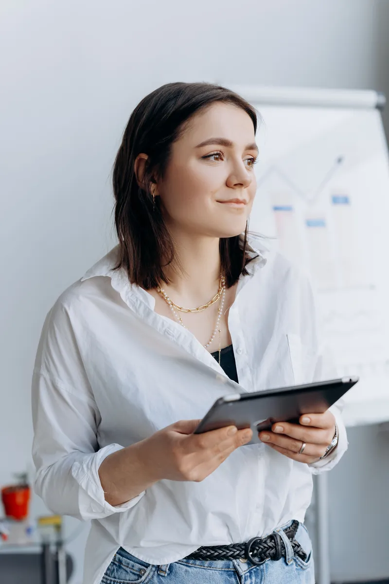 A woman in a white shirt holds a tablet in front of a flip chart showing a line graph and bar chart