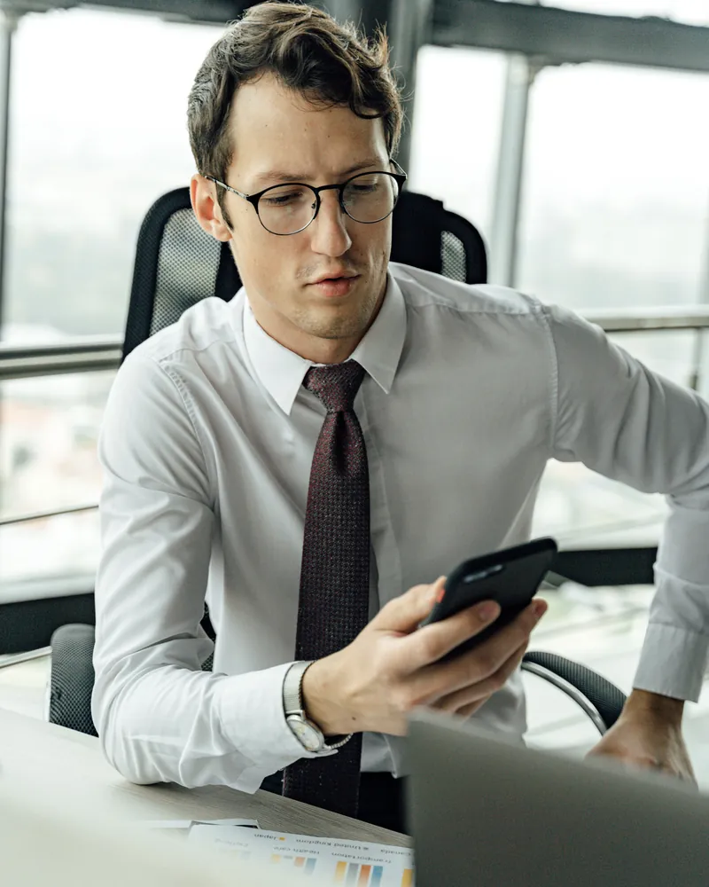 A man in a white shirt and dark tie looks at his phone at an office desk, beside a laptop and a printed bar chart