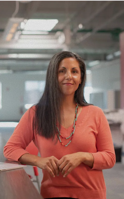 A woman with long dark hair and a turquoise and coral beaded necklace in a coral top in an open-plan office