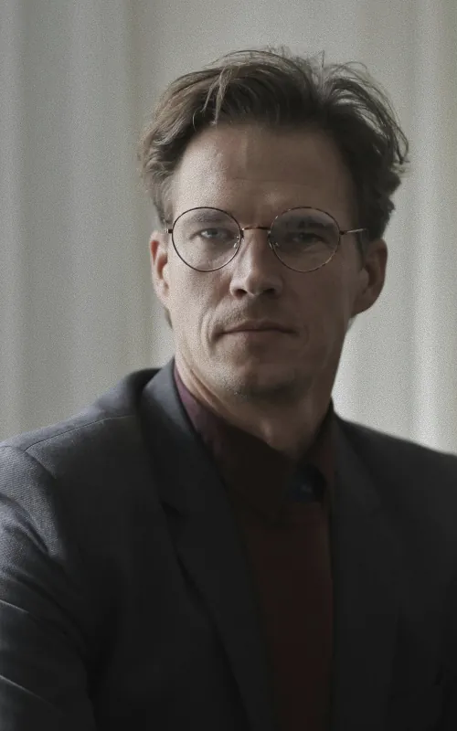 A man with wavy light brown hair and round glasses in a grey blazer over a dark red shirt against a pale curtain