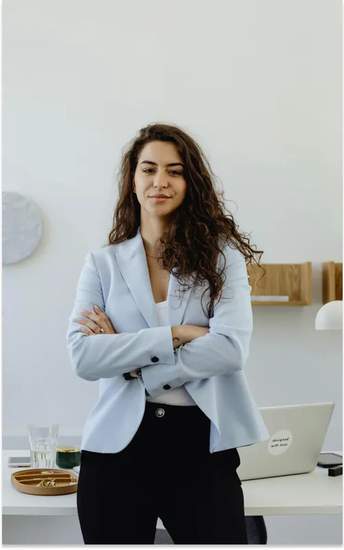 A woman with long curly dark hair in a pale blue blazer, arms folded, standing at a white desk