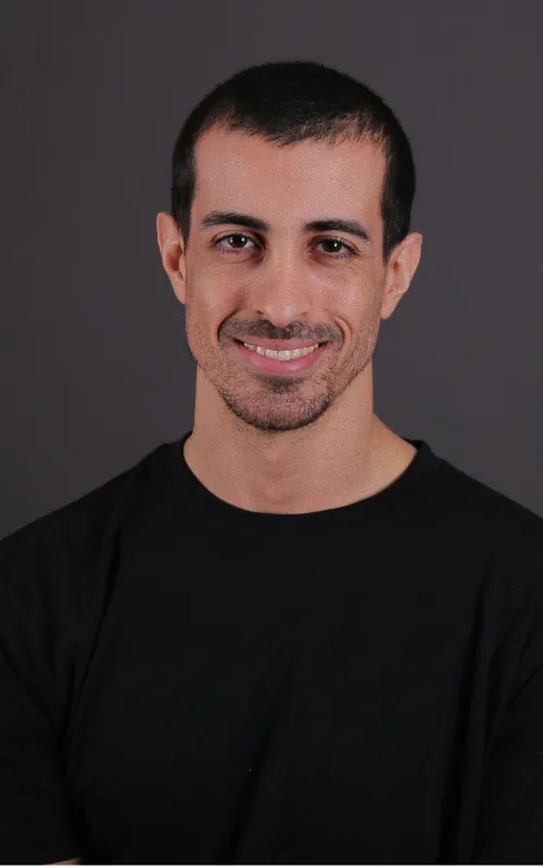 Un homme aux cheveux courts bruns et à la barbe naissante, en t-shirt noir, sur un fond de studio gris foncé