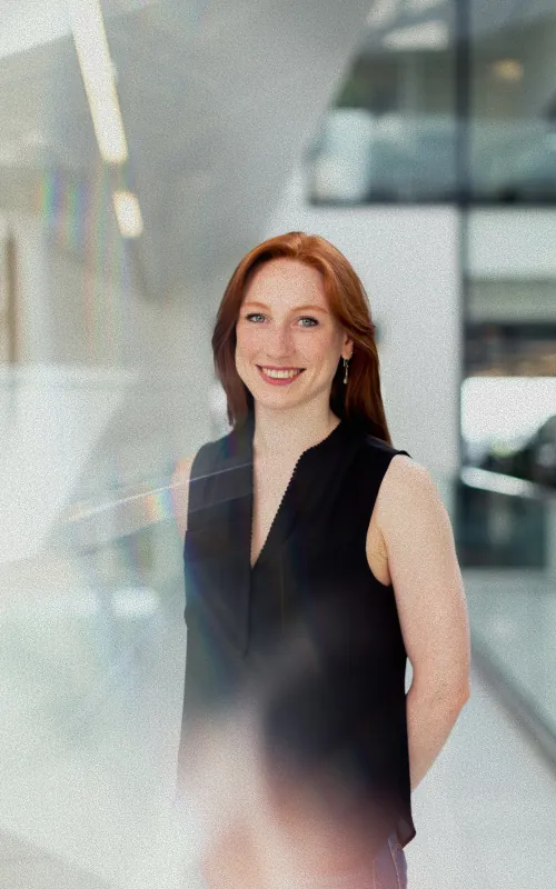 A woman with long red hair in a black sleeveless top standing in a bright modern glass interior