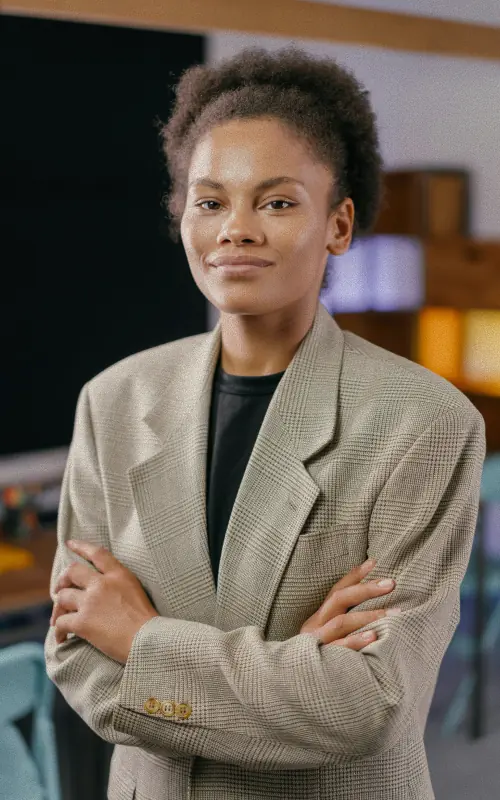 A woman with her hair gathered up, in an oversized beige checked blazer over a black t-shirt, arms folded