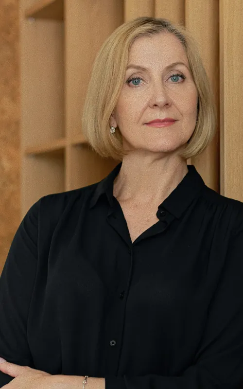 A woman with a chin-length blonde bob in a black shirt, arms folded, in front of a pale wooden slatted wall