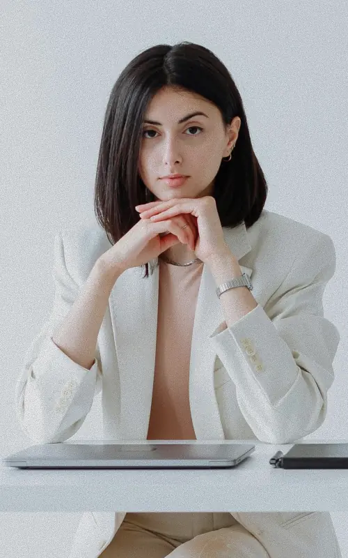 A woman with a dark bob in an oversized cream blazer, seated at a white desk with her chin resting on her clasped hands