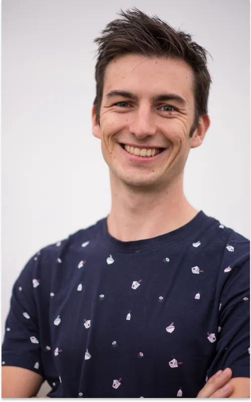 Un jeune homme souriant en t-shirt bleu marine imprimé de petits motifs, bras croisés, sur fond gris clair