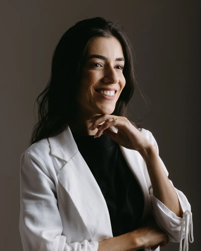 Retrato de estudio de una mujer con blazer blanco sobre fondo oscuro, sonriendo con la mano apoyada en la barbilla