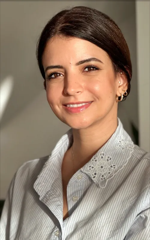 A smiling woman with dark hair pulled back, in a pale blue striped shirt with an embroidered collar, in a warm interior