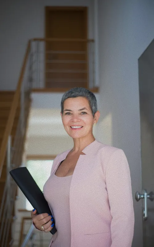 A woman with short grey hair in a pink blazer holding a folder, standing in a bright interior with a staircase behind her
