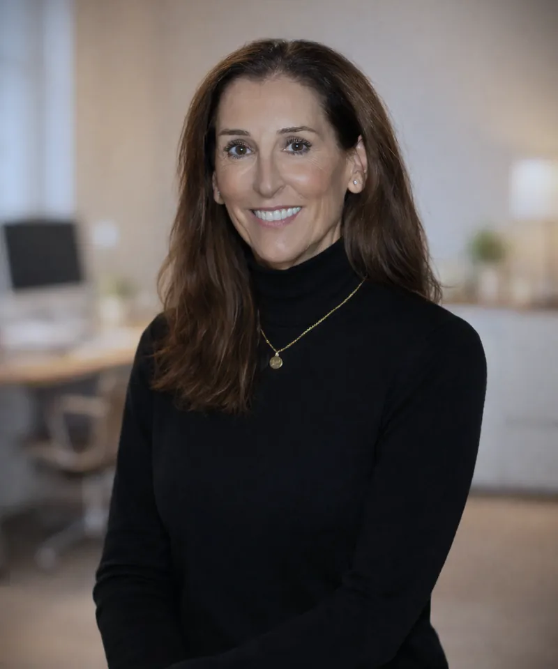 A smiling woman with long brown hair in a black turtleneck and a small gold pendant, in a bright office