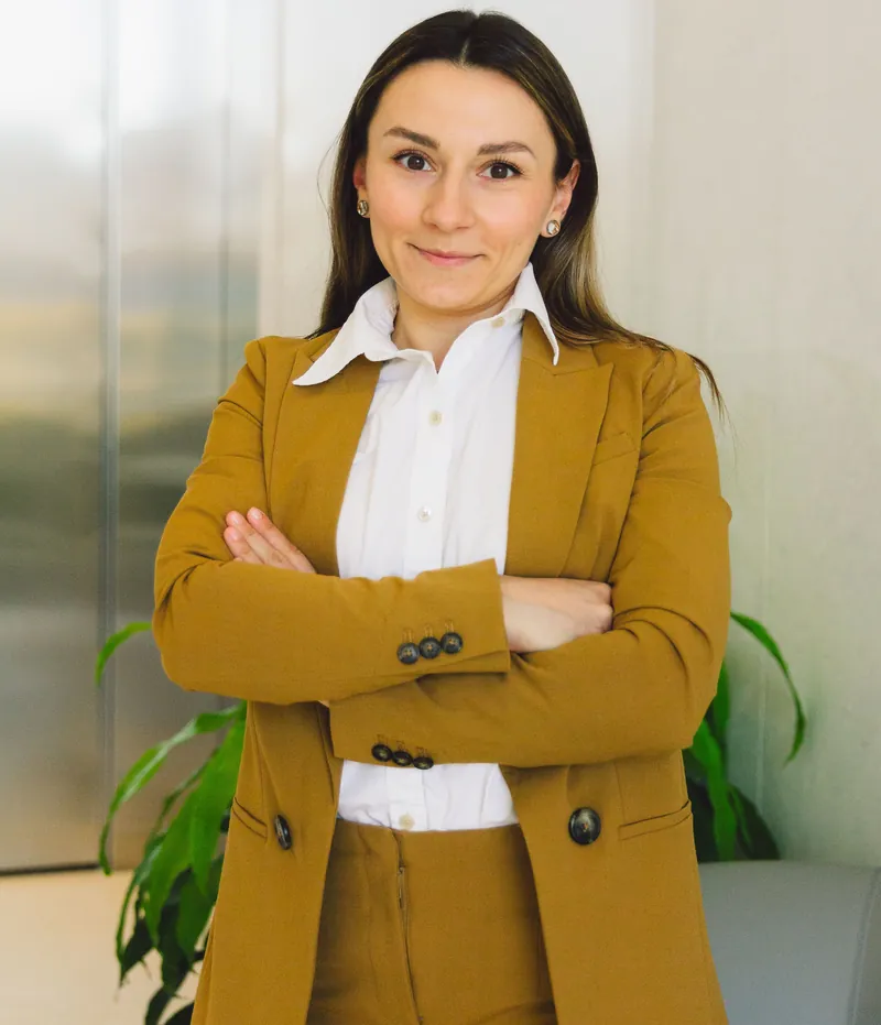 Retrato de una mujer con traje color mostaza y camisa blanca, de pie y con los brazos cruzados, en una oficina de paredes acristaladas